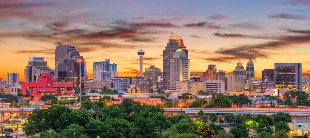 San Antonio city skyline at sunset, featuring tall buildings, the Tower Life Building, and a colorful sky with orange and purple clouds above a foreground of green trees.