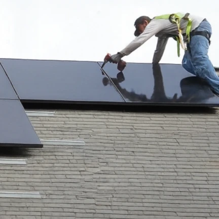A worker wearing a safety harness installs black solar panels on a sloped, shingled roof against a light sky background.