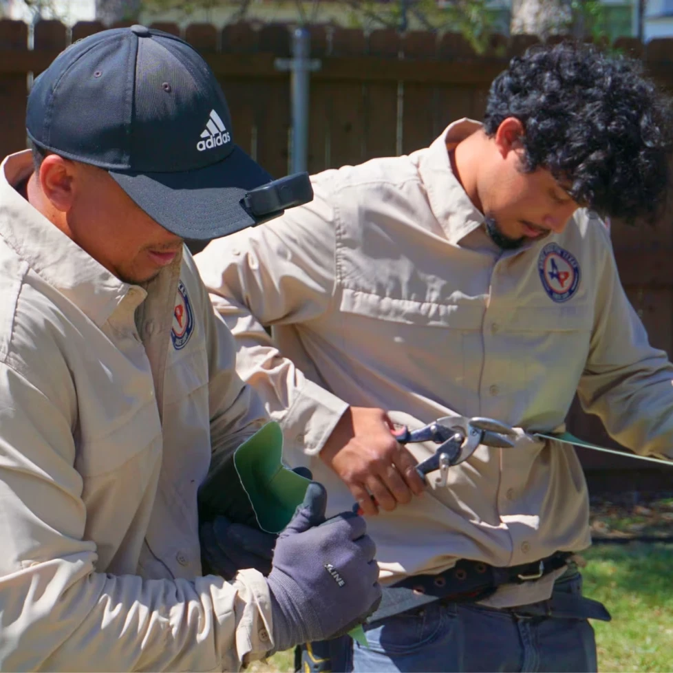 Two men wearing beige uniforms and gloves work outdoors with electrical tools and wires. Both focus intently on their tasks; one holds wire and pliers, the other appears to wear a head-mounted camera or light.