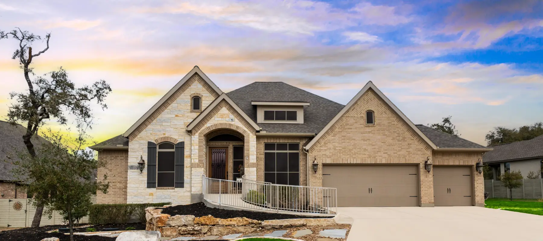 A modern single-story house with a stone and brick exterior, large windows, an arched entryway, a ramp leading to the front door, and a double garage, set against a colorful sunset sky.