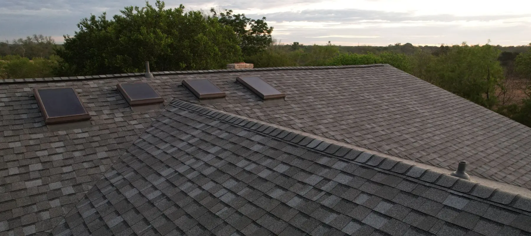 A rooftop with gray asphalt shingles features four rectangular skylights and a ridge vent, surrounded by trees with a cloudy sky in the background.