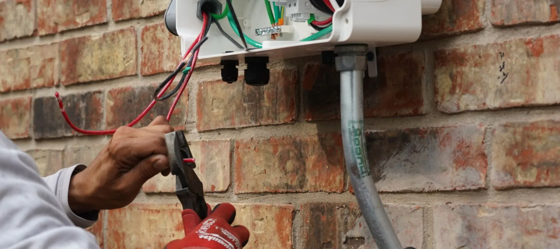A person wearing red gloves uses pliers to work on electrical wiring connected to a white junction box mounted on a brick wall. Exposed wires and conduits are visible coming out of the box.