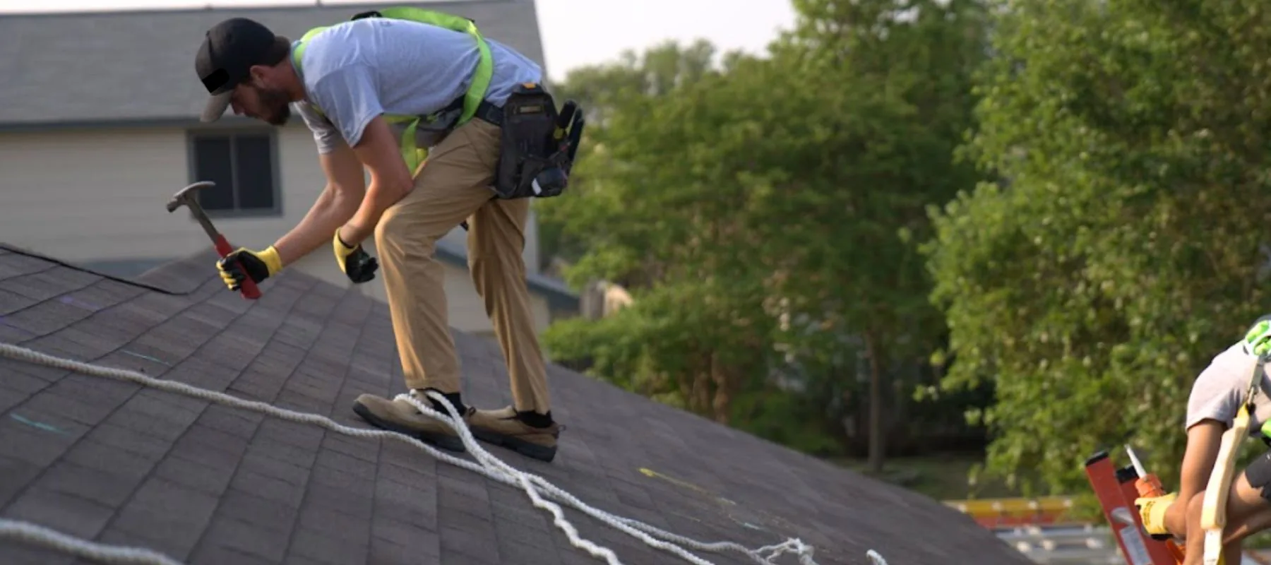 A person wearing a safety harness uses a hammer while working on a sloped roof, with safety ropes secured and trees and houses visible in the background.