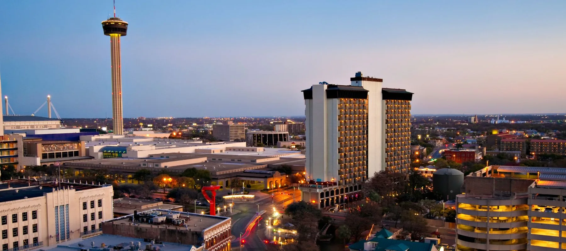A cityscape of downtown San Antonio at dusk, featuring the Tower of the Americas, tall modern buildings, illuminated streets, and a soft blue sky.
