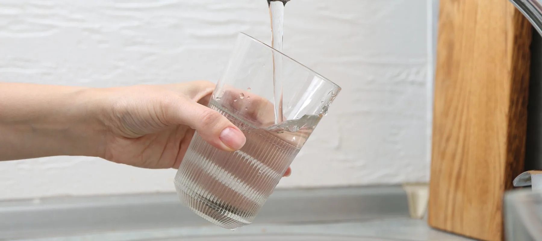 A hand holds a clear, ribbed glass under a running kitchen faucet, filling it with water. The background shows a white textured wall and part of a wooden object.