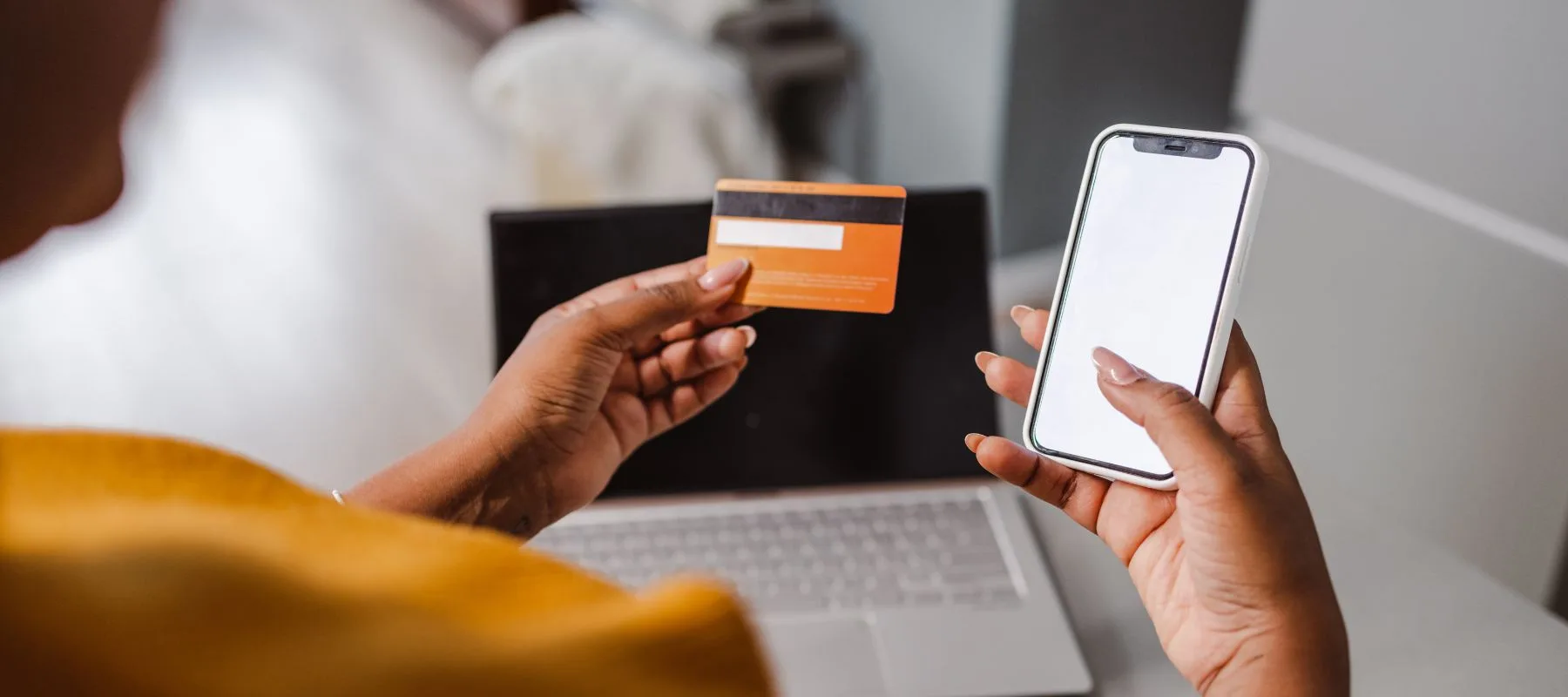 A person holds a credit card in one hand and a smartphone with a blank screen in the other, sitting at a desk with an open laptop in the background.