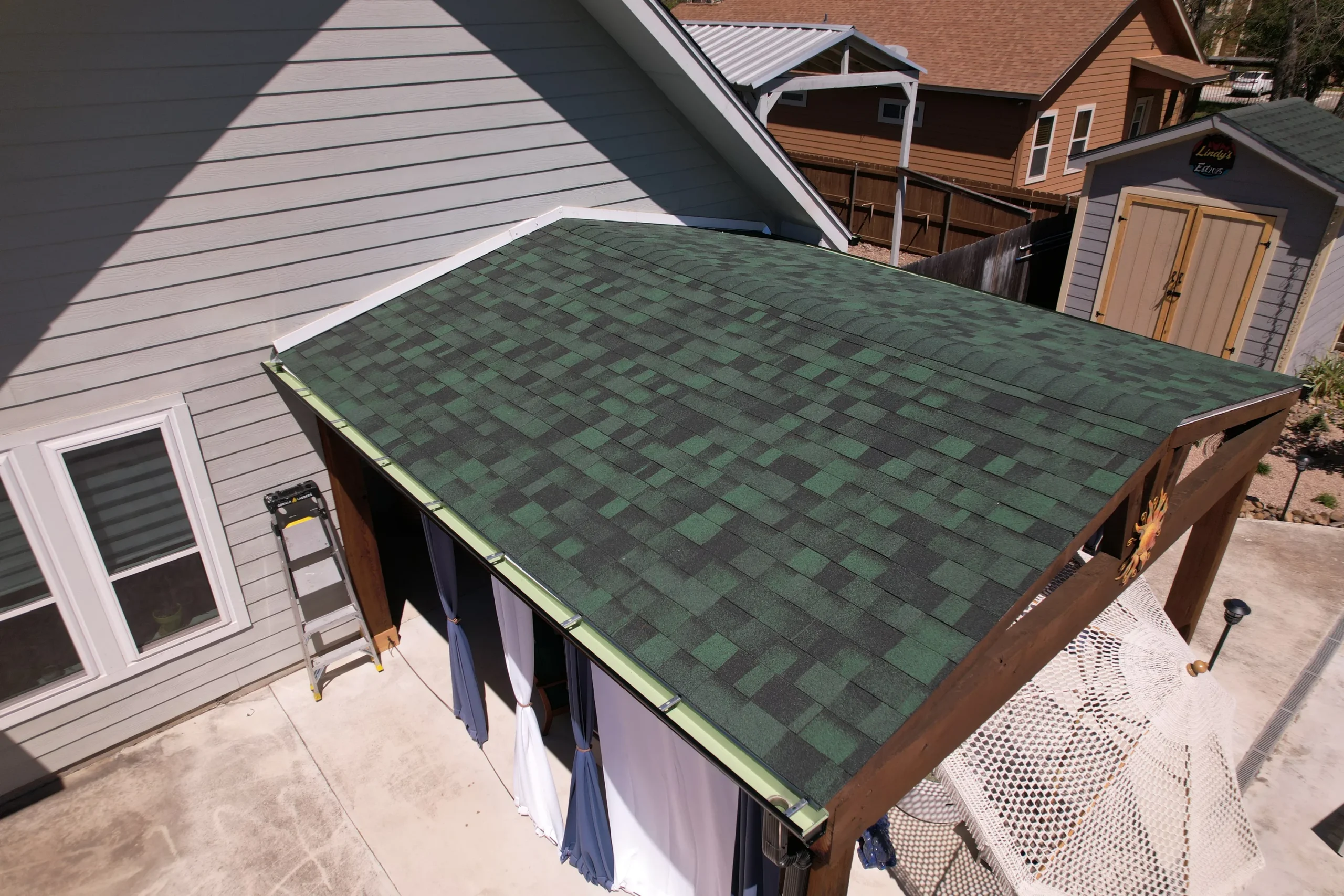 A green-shingled roof covers a wooden patio structure beside a gray house. White and blue curtains hang from the patio, and a ladder leans against the house. Nearby are a shed and another house.