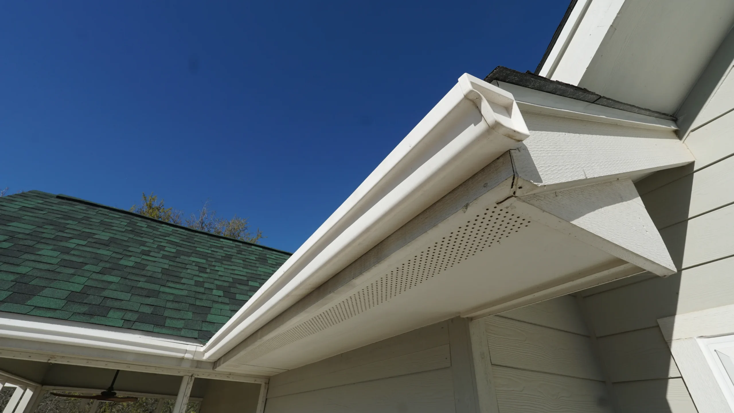 Close-up view of a white rain gutter attached to the edge of a house roof with green shingles, set against a clear blue sky.