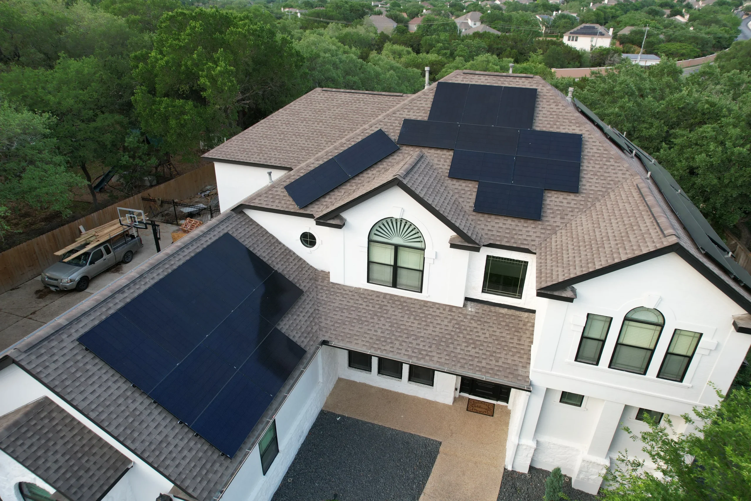 A large house with multiple solar panels installed on its roof, surrounded by trees and greenery, with a driveway and a parked vehicle visible.