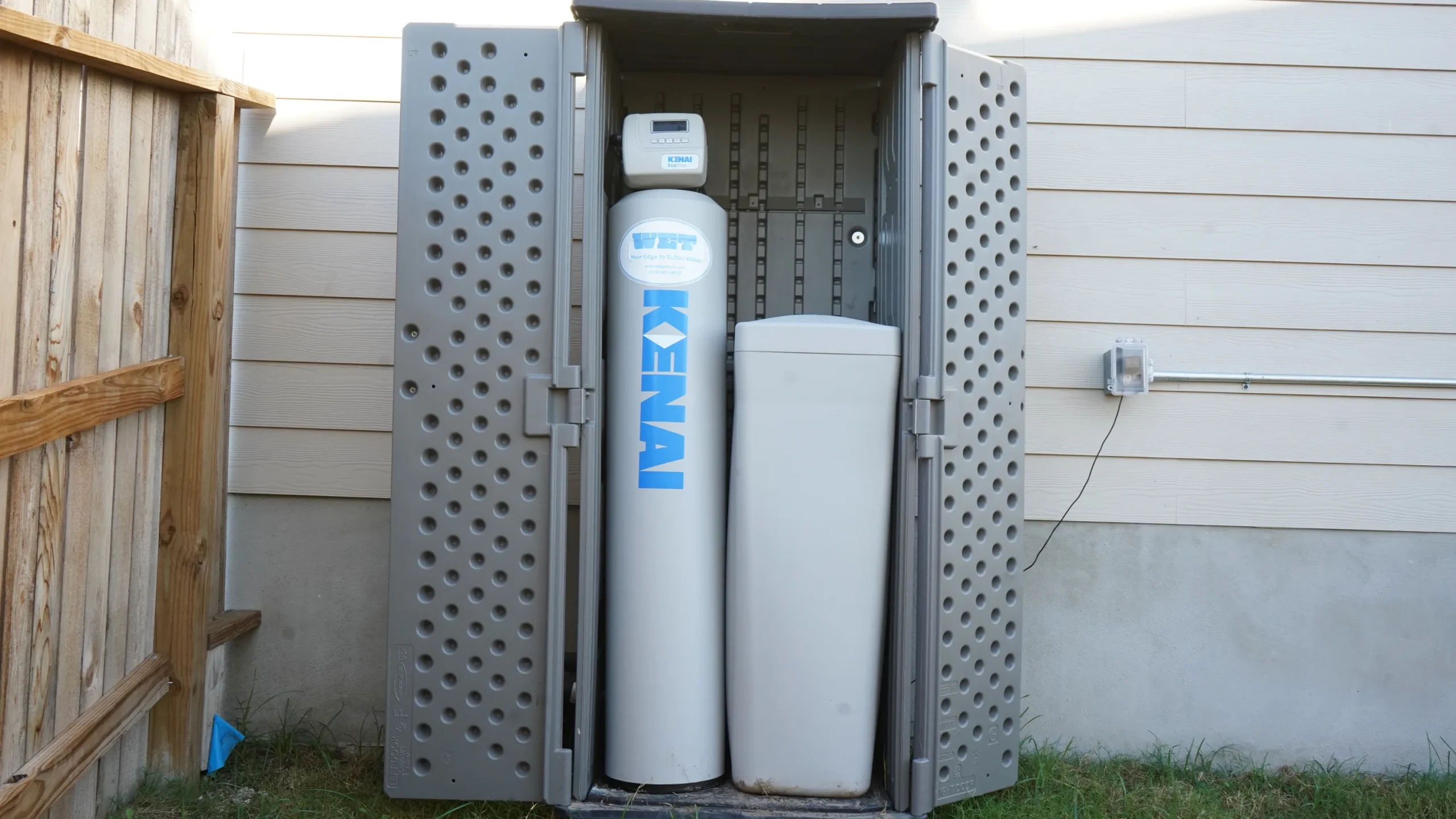 A gray outdoor cabinet with perforated doors stands open, revealing a Kenai water softener system with two tall, cylindrical tanks positioned against a house’s exterior wall.
